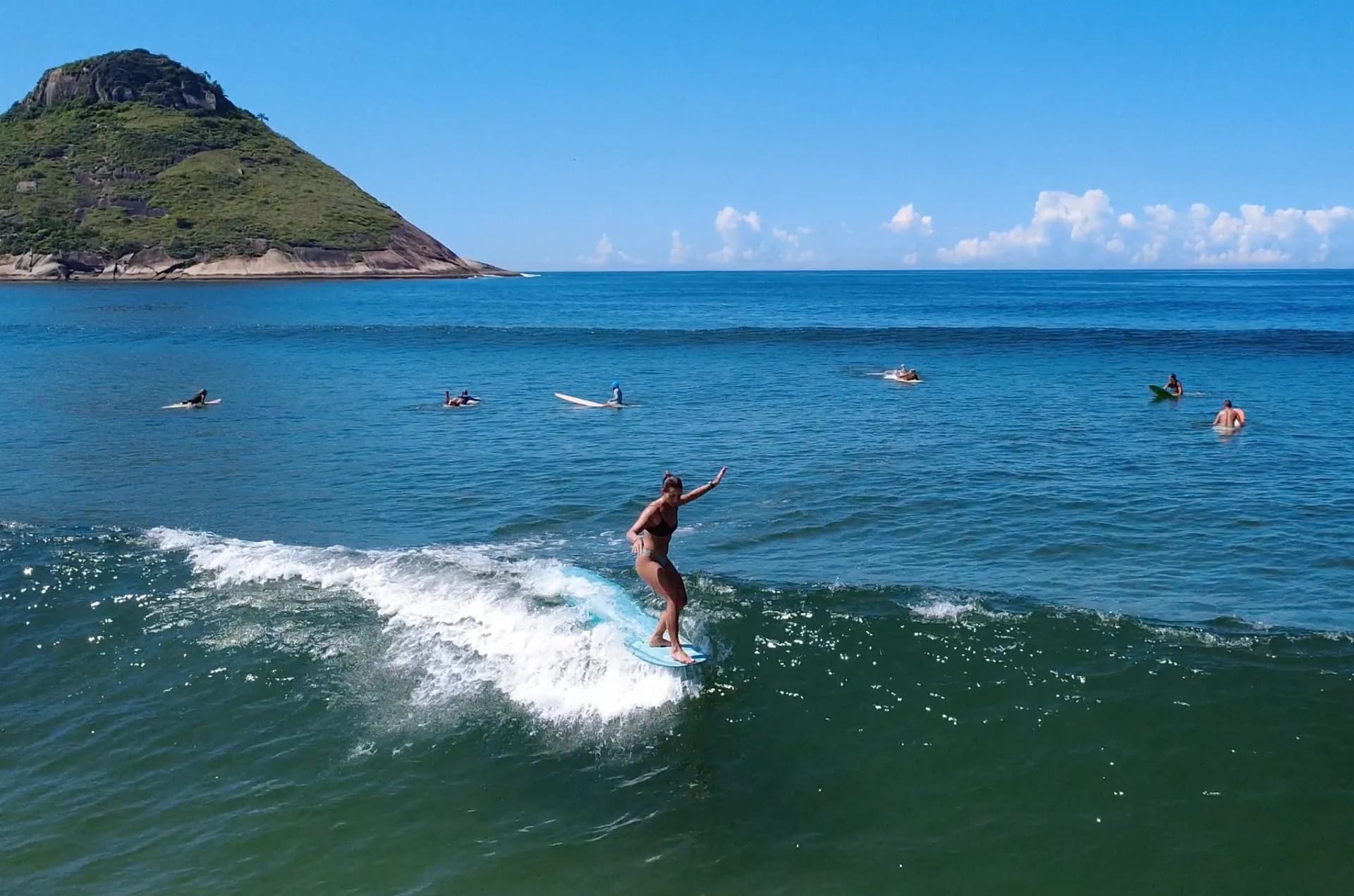 Surfista em uma prancha turquesa numa onda, com outros surfistas e uma ilha verde ao fundo